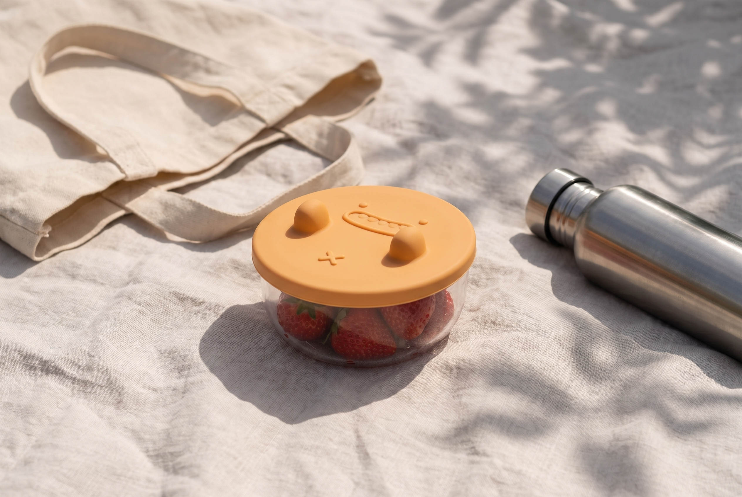 The sealed snack pot of strawberries on a picnic blanket beside a canvas tote
